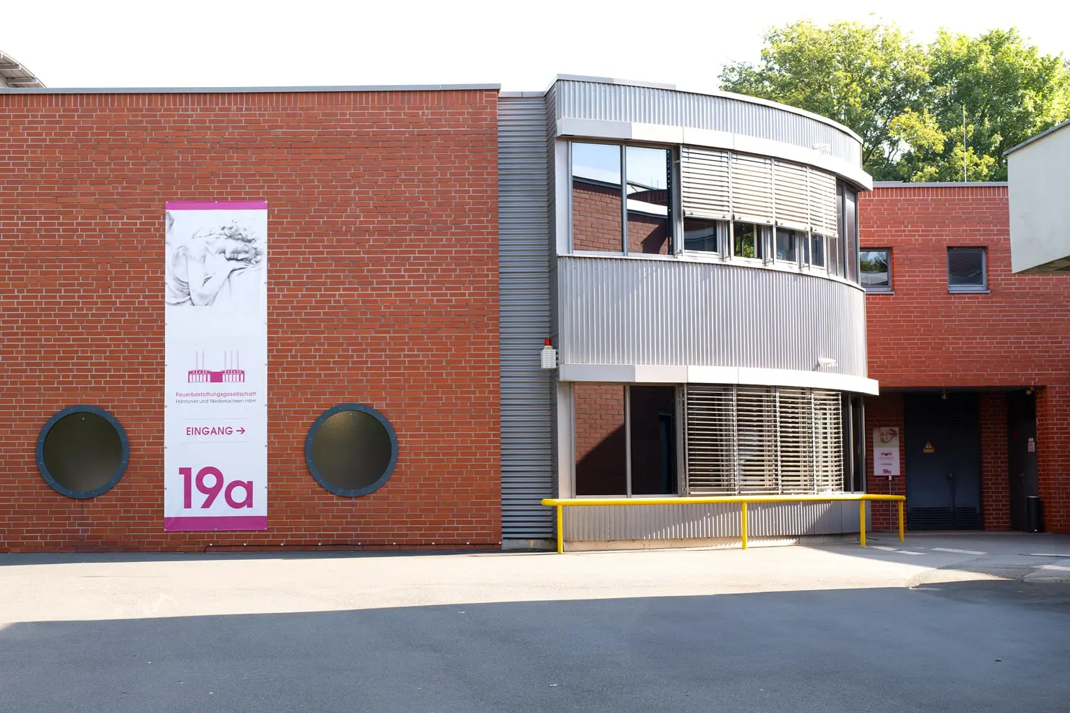 Eingangsbereich der Feuerbestattungsgesellschaft Hannover mit Ziegelwand, modernem Anbau und Schild 'Eingang 19a'.