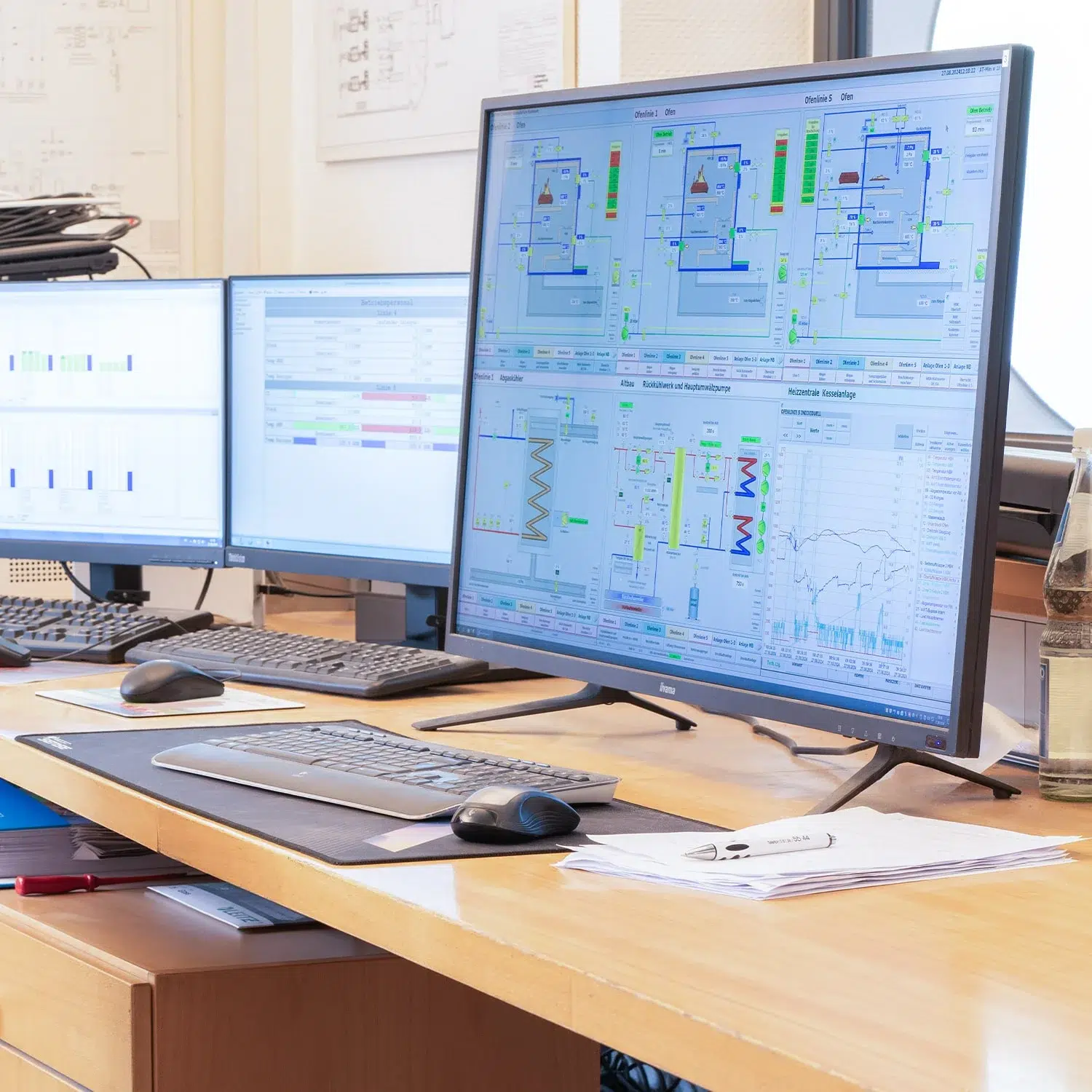 Schreibtisch mit drei Bildschirmen, die Prozessleitsysteme und Anlagendaten darstellen.