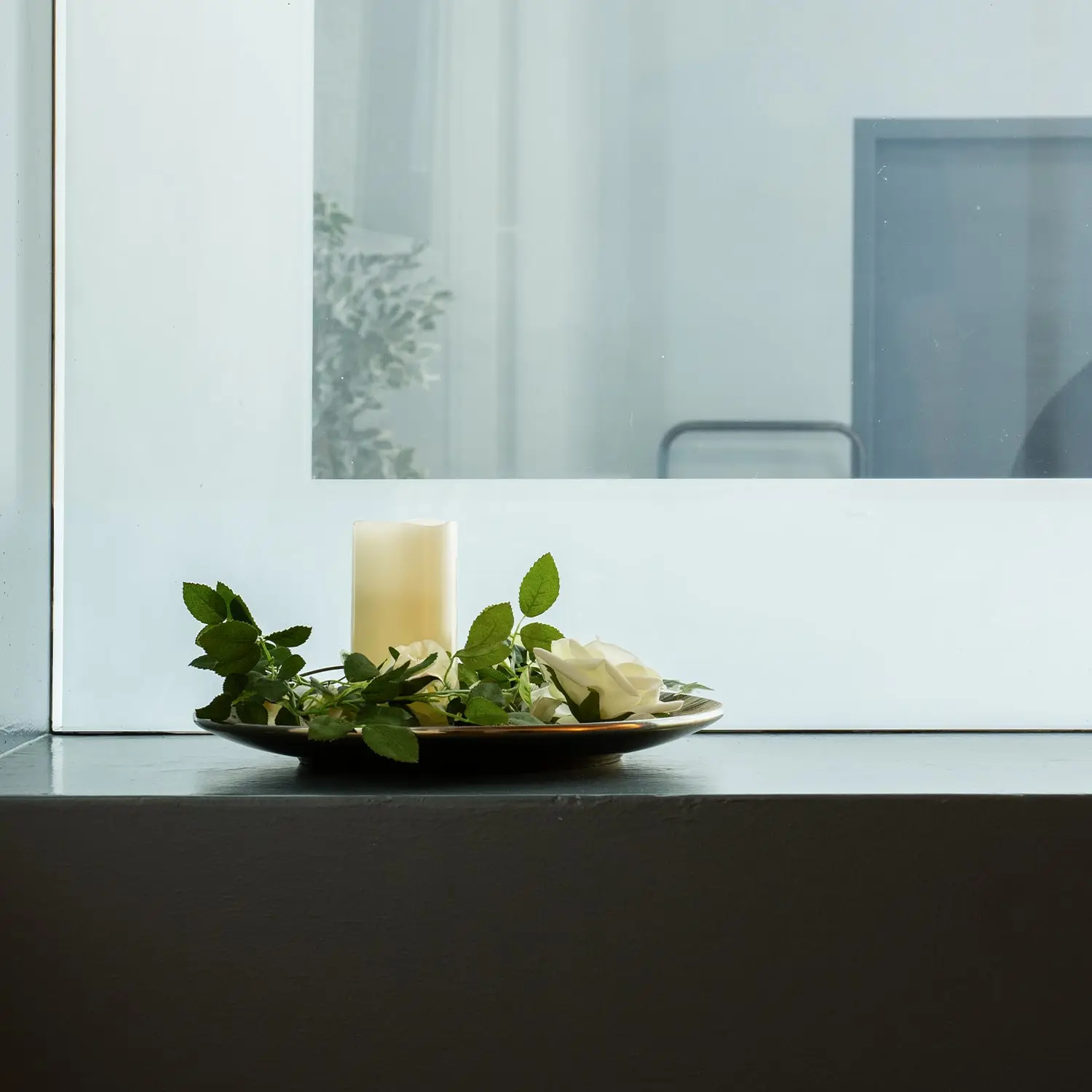 Cremefarbene Kerze, weiße Rosen und grünes Blattwerk auf einem Dekoteller.