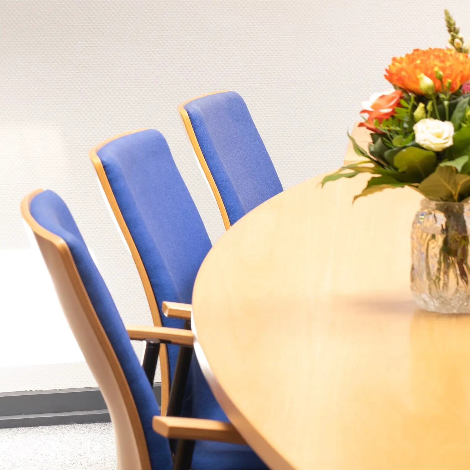 Ein heller Holztisch mit blumen und blauen stühlen in einem besprechungsraum.